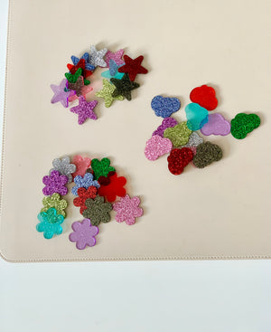 Assorted glittery plastic shapes including stars, flowers, hearts, and clouds scattered on a beige surface