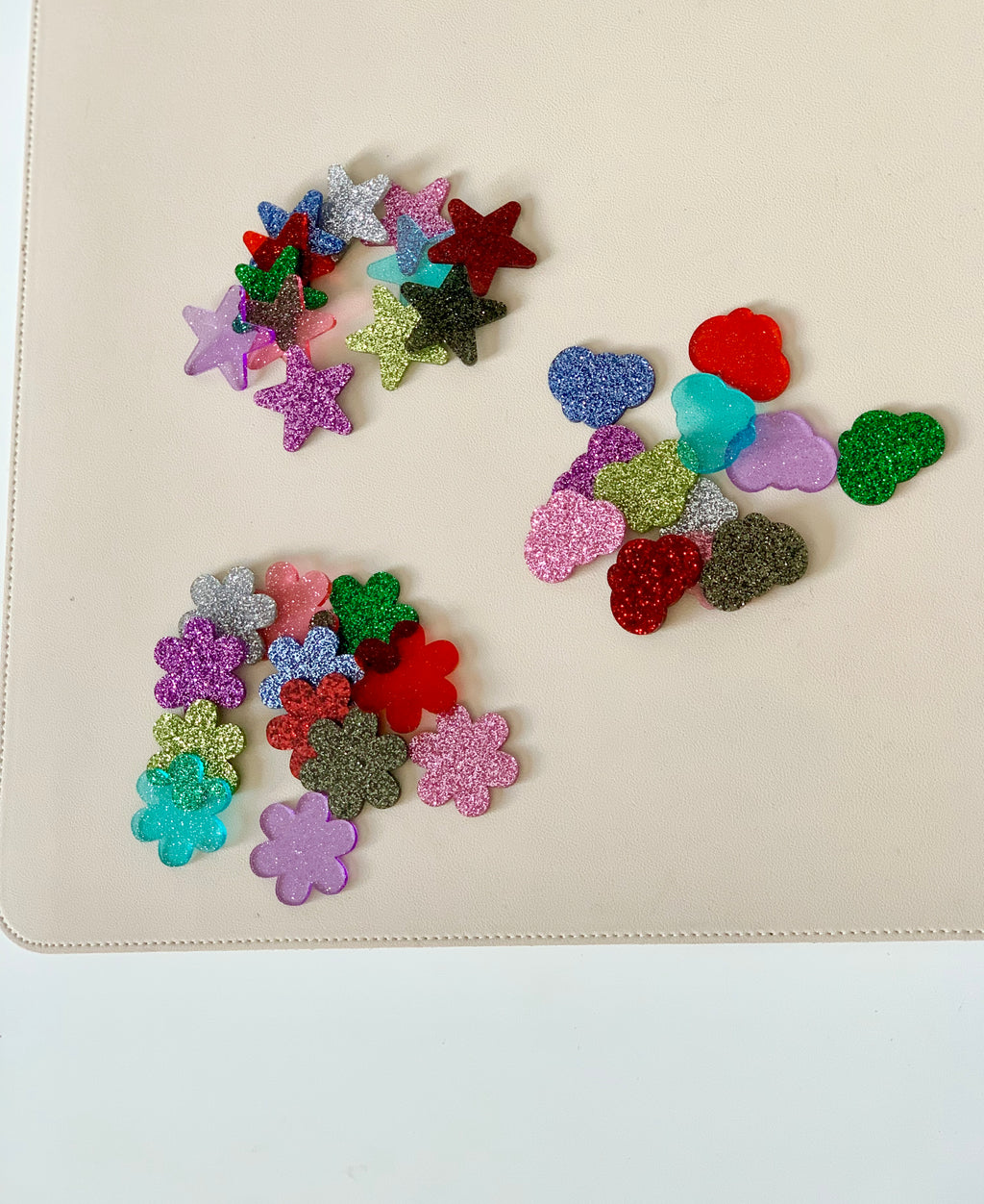 Assorted glittery plastic shapes including stars, flowers, hearts, and clouds scattered on a beige surface