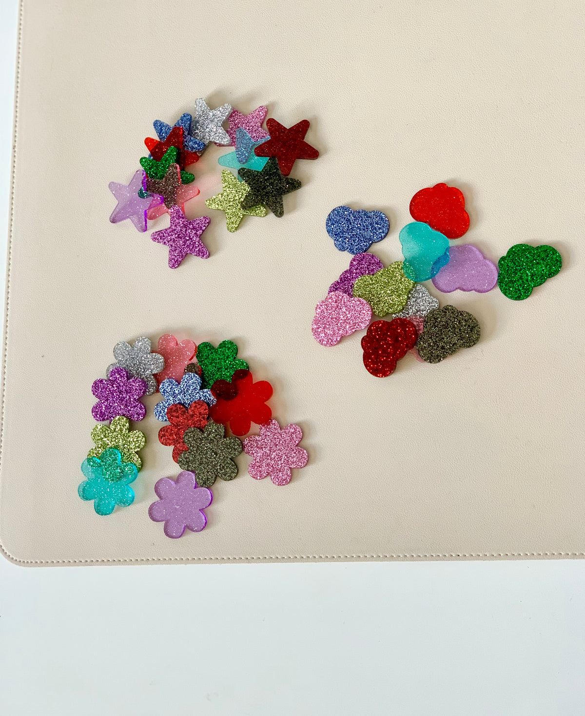 Assorted glittery plastic shapes including stars, flowers, hearts, and clouds scattered on a beige surface