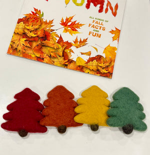 Four felt pine tree shapes in red, orange, yellow, and green with brown trunks on a white surface next to a fall-themed book