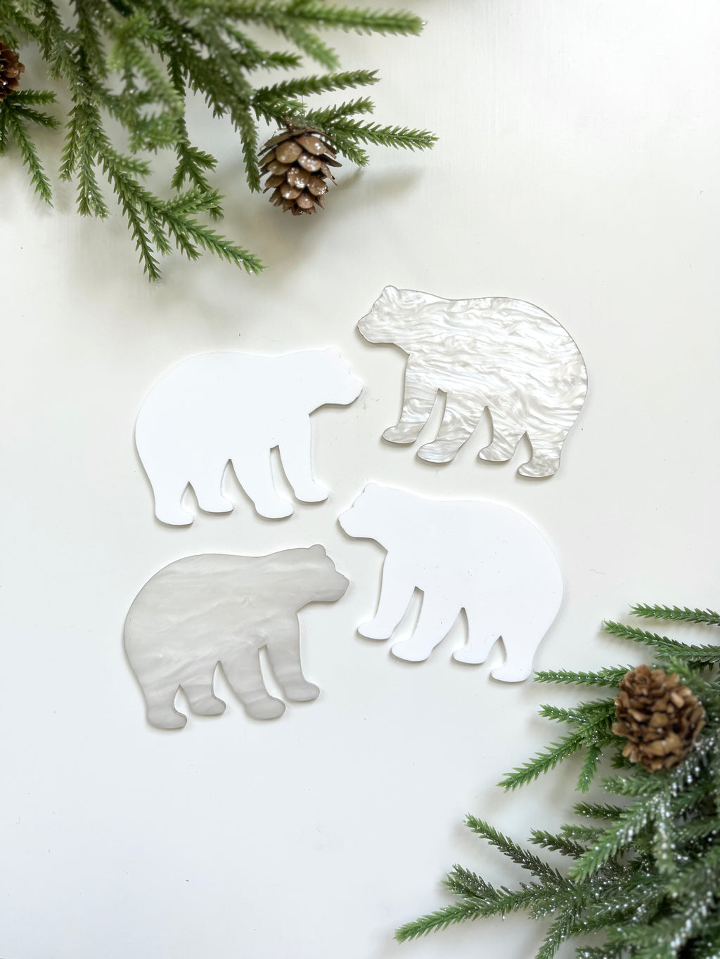 Four white and pearlescent bear-shaped cutouts on white surface with pine branches and pinecones around edges