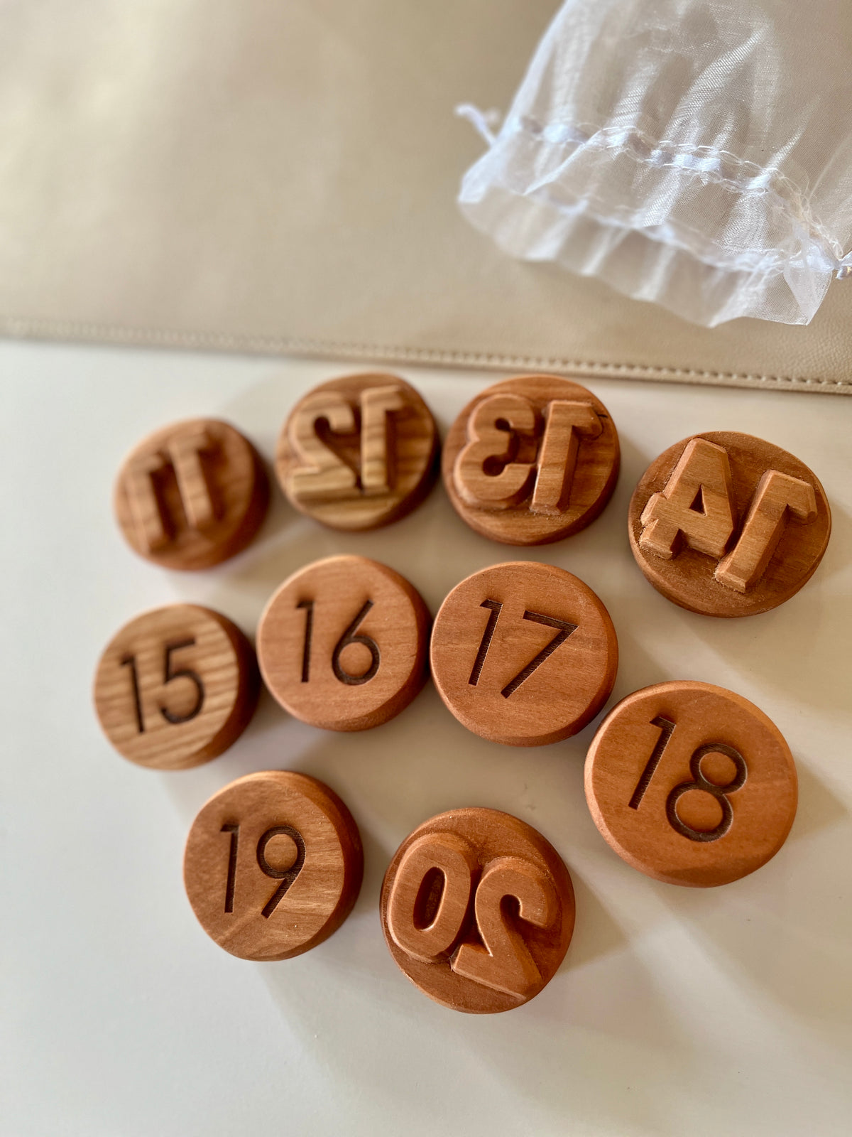 Round wooden tokens numbered 11 to 20 with carved and raised number styles on a light surface next to a sheer pouch