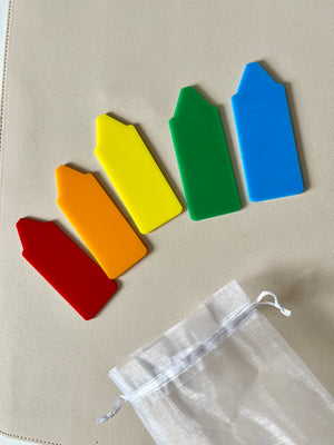 Five brightly colored plastic plant markers in red, orange, yellow, green, and blue laid out on a light surface with a white drawstring bag nearby