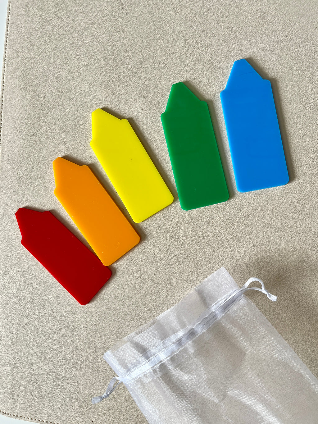 Five brightly colored plastic plant markers in red, orange, yellow, green, and blue laid out on a light surface with a white drawstring bag nearby