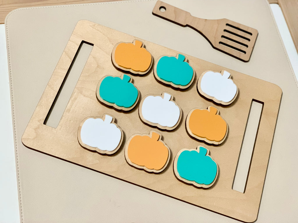 Wooden cookie sheet with orange, teal, and white pumpkin-shaped cookies and a wooden spatula on beige surface