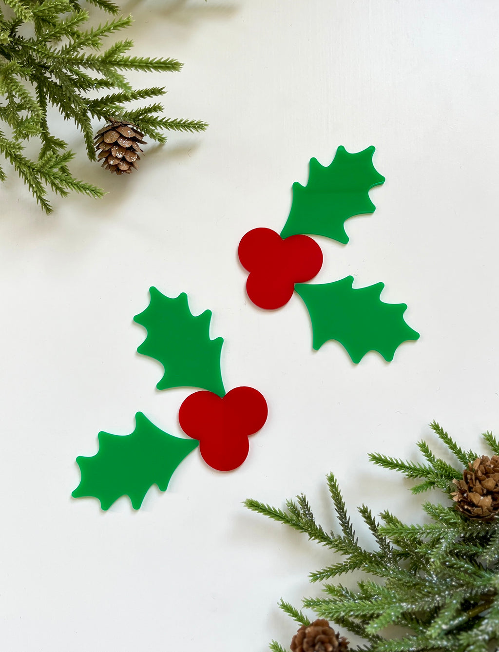 Two green holly leaf cutouts with red berry clusters surrounded by pine branches and pinecones on white background