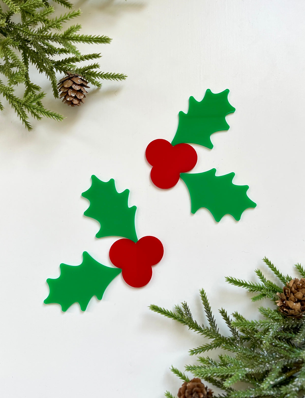 Two green holly leaf cutouts with red berry clusters surrounded by pine branches and pinecones on white background
