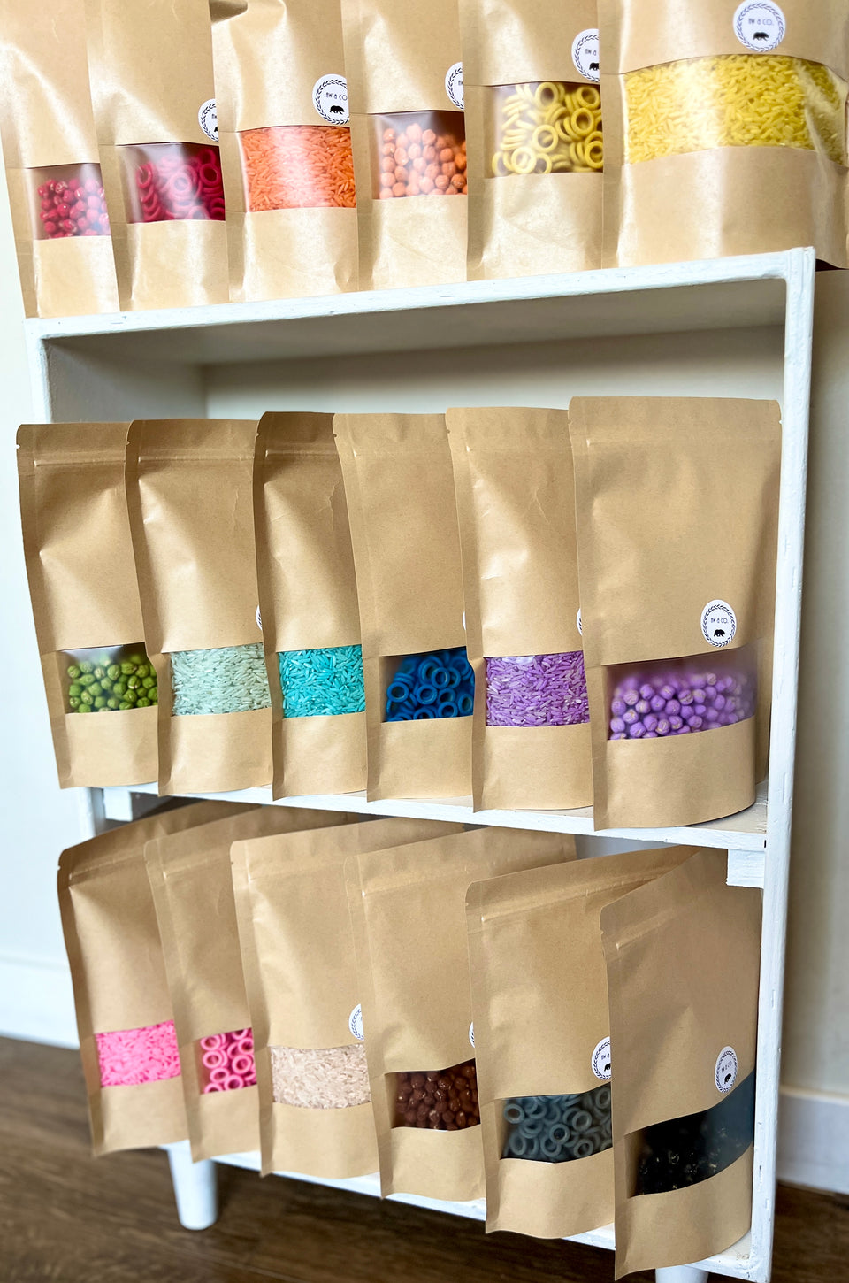 Multiple brown kraft paper bags filled with assorted brightly colored sensory filler types arranged on white wooden shelves