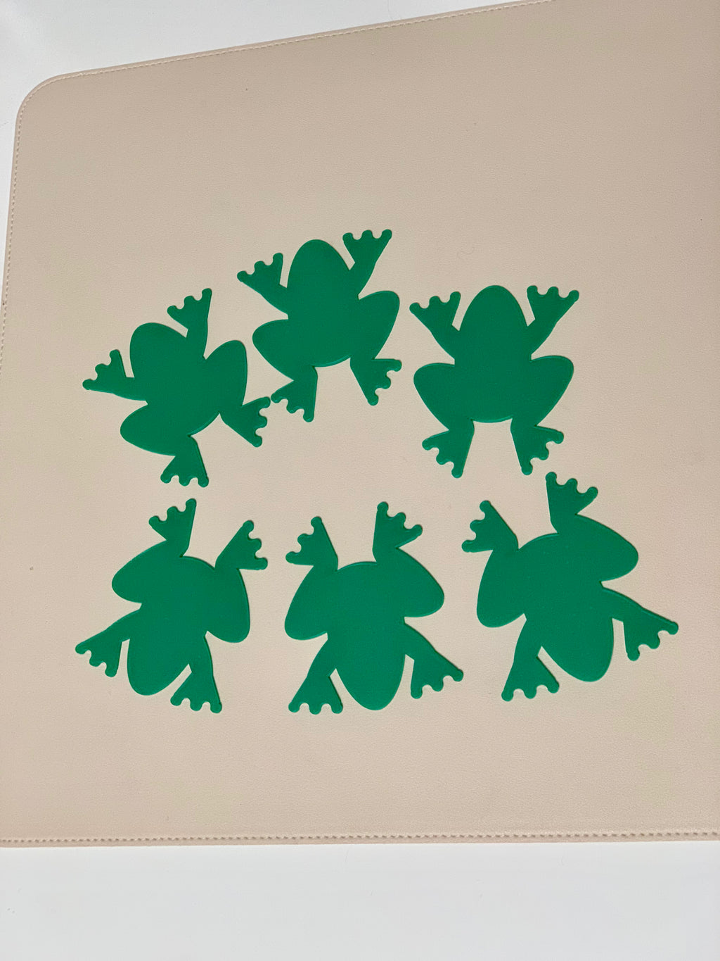 Six solid green frog-shaped cutouts arranged in a circular pattern on a beige surface