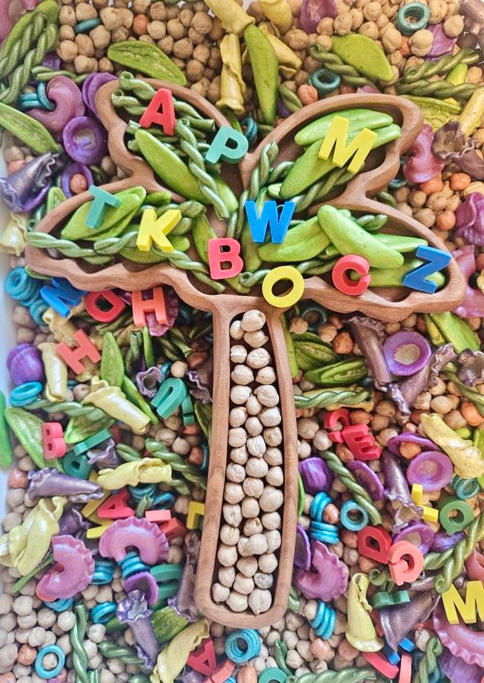 Wooden tree-shaped tray filled with chickpeas and green pasta surrounded by colorful alphabet letters and dried pasta pieces