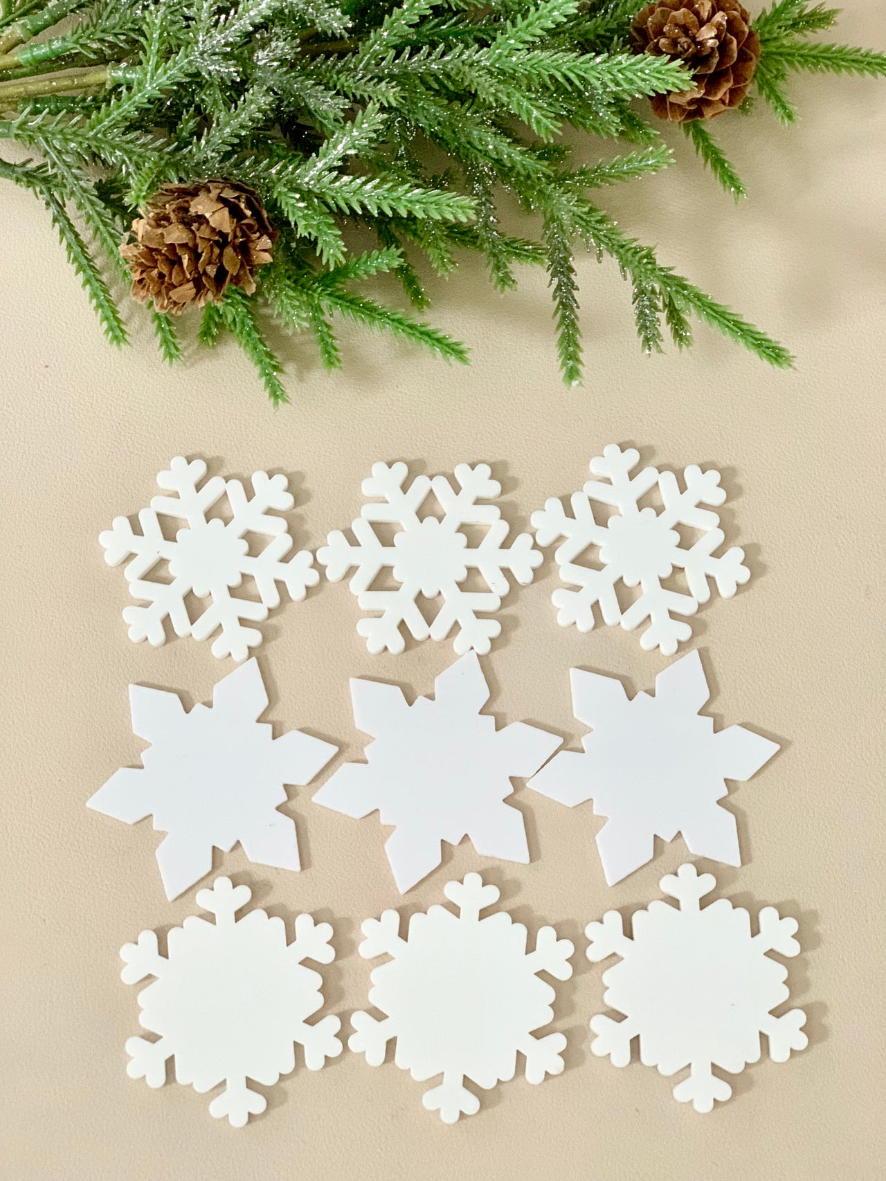 Nine white snowflake-shaped cutouts in three distinct designs arranged on beige surface with pine branches and cones above