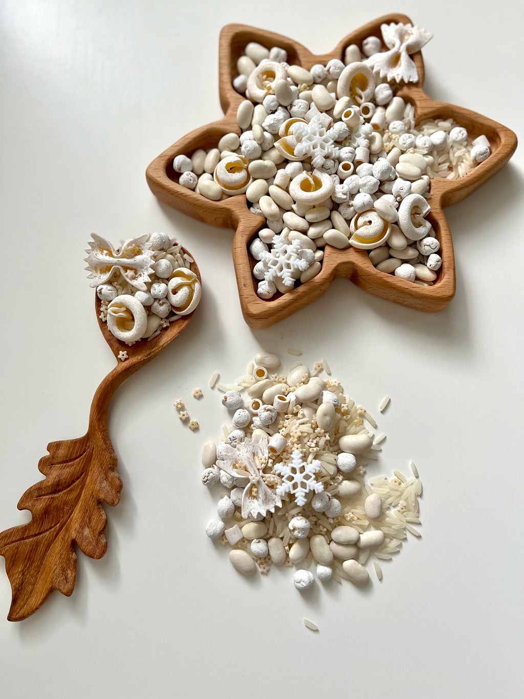 Star-shaped wooden tray and leaf-shaped spoon filled with white beans, rice, pasta, and decorative snowflakes on white surface