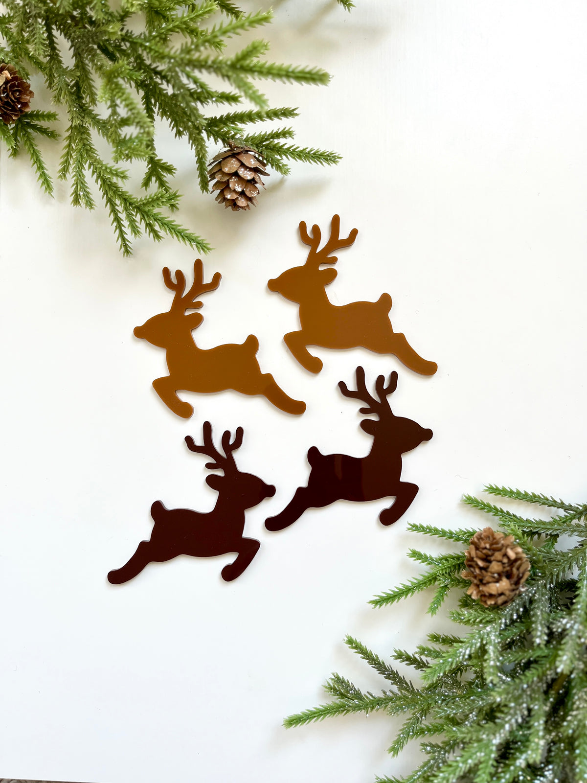 Four brown reindeer cutouts arranged on white background with green pine branches and pinecones at corners
