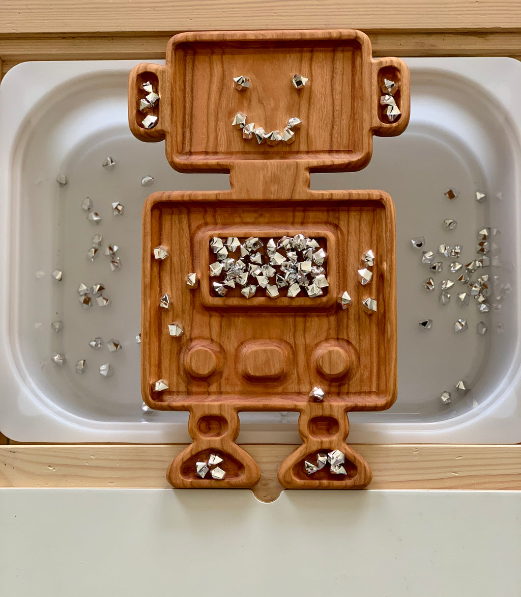 Wooden robot-shaped sensory tray with silver confetti pieces arranged on a white surface background