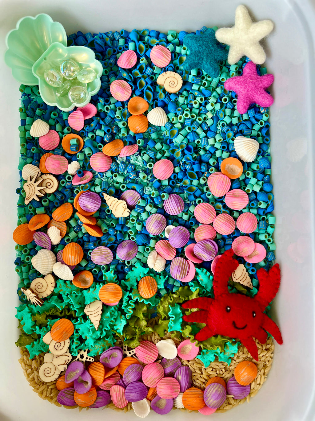 Sensory bin with blue pasta, colorful wooden shells, felt starfish, clear beads, and a red felt crab figure.