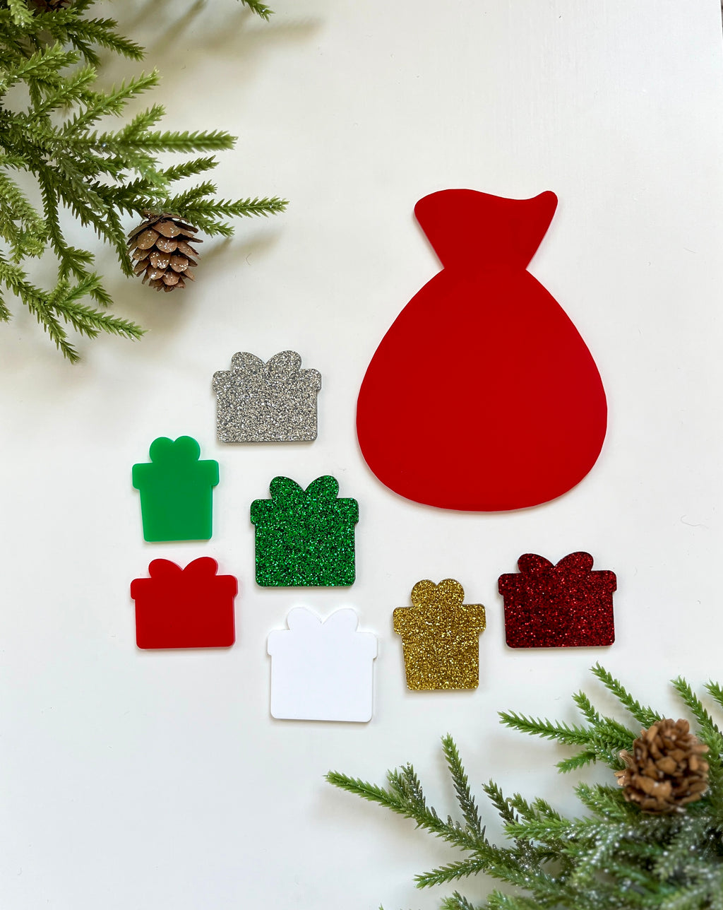 Red Santa sack cutout surrounded by seven glittery and matte gift-shaped cutouts in red, green, gold, silver, and white on white background with pine branches and cones