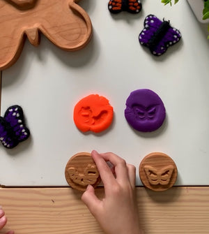 Child’s hand pressing wooden butterfly stamps creating orange and purple clay impressions on white table with felted butterfly toys nearby