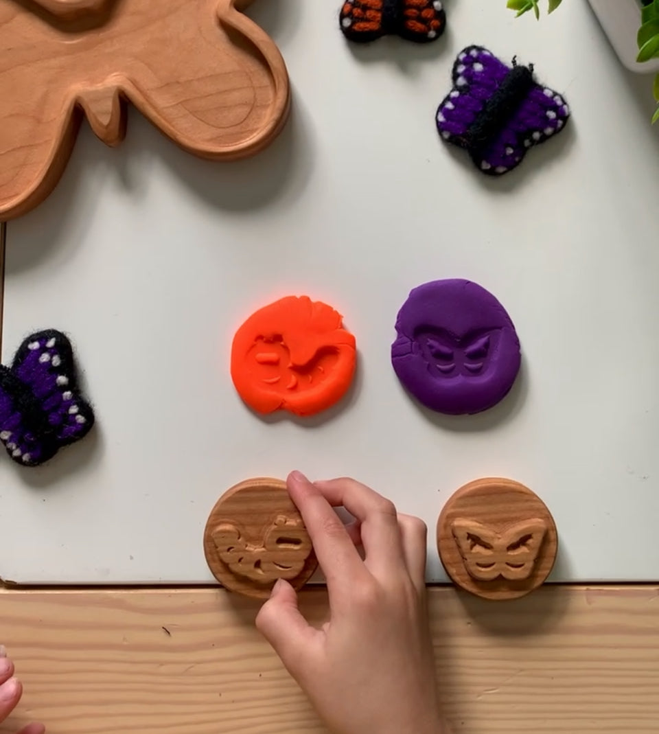 Child’s hand pressing wooden butterfly stamps creating orange and purple clay impressions on white table with felted butterfly toys nearby