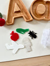 Five colorful acrylic shapes including airplane, apple, crocodile, ant, and key next to wooden letter trays on white surface