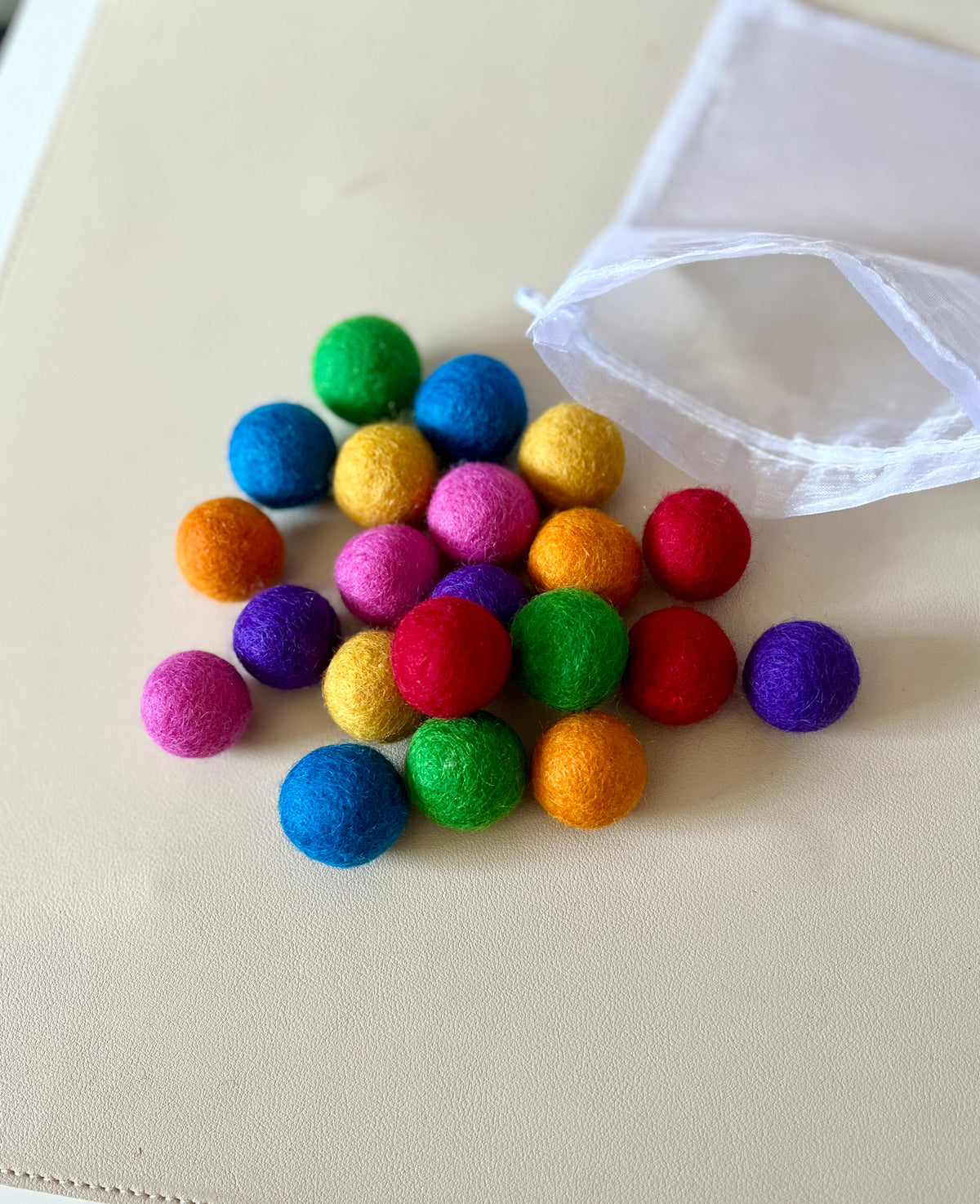 Pile of colorful felt balls in rainbow shades spilling out of white mesh bag on light surface