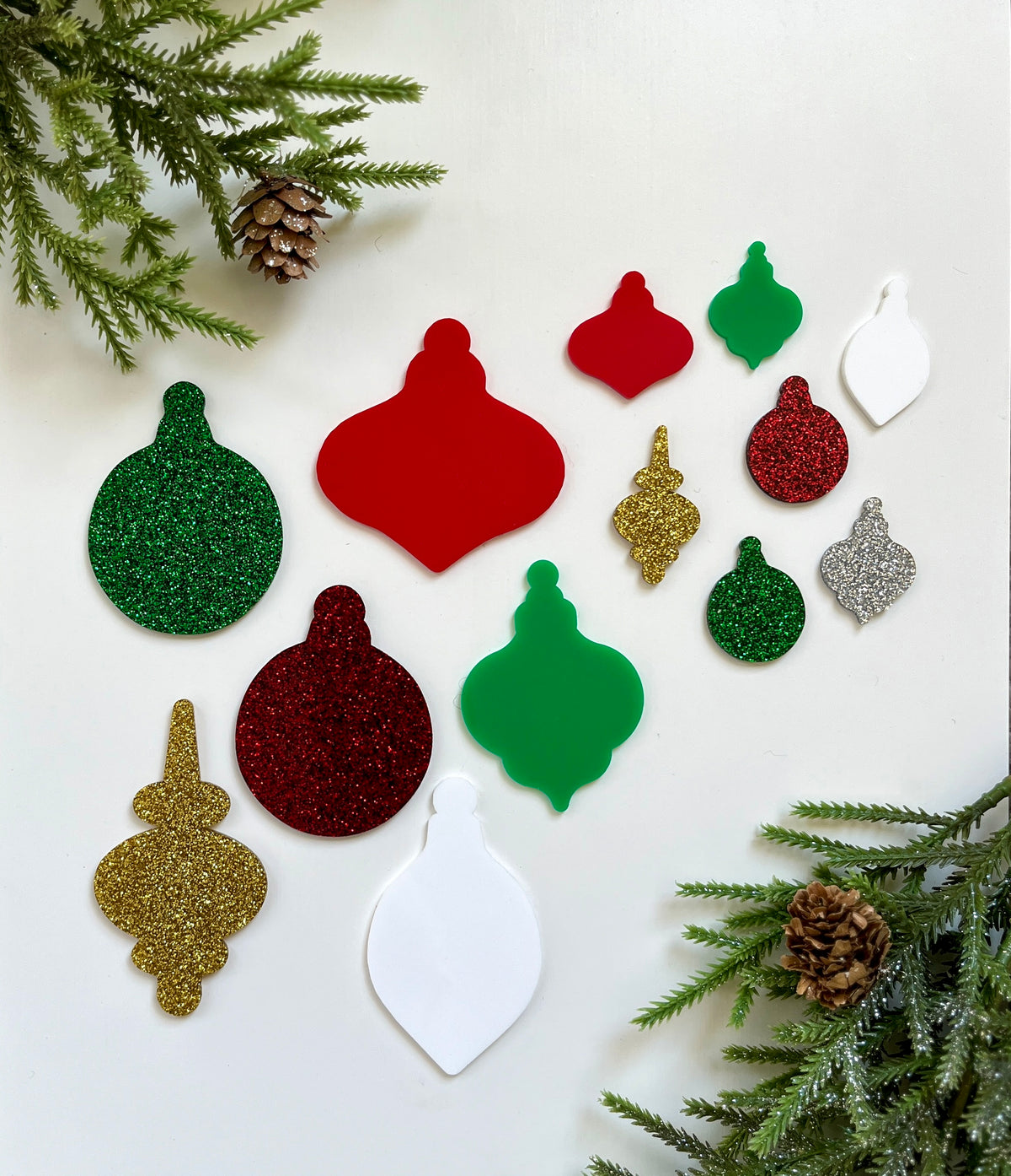 Assorted glittery and matte red, green, gold, silver, and white Christmas ornament cutouts on white background with pine branches and cones