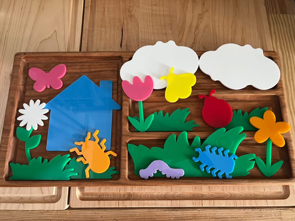 Wooden tray with colorful plastic garden shapes including flowers, bugs, clouds, butterfly, house, and grass pieces