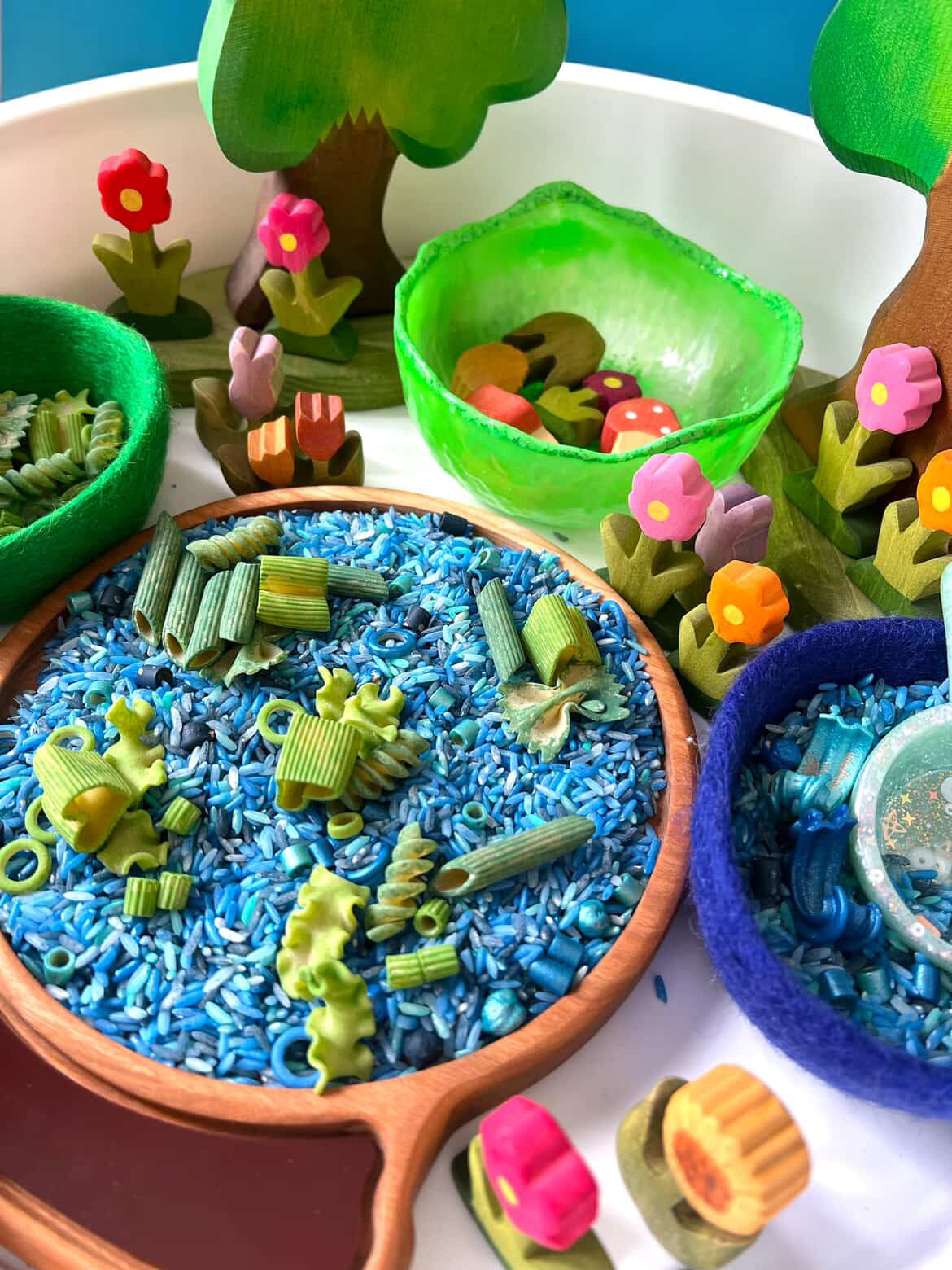 Colorful sensory play setup with blue rice, green pasta, wooden trees, and felt bowls on a white tray