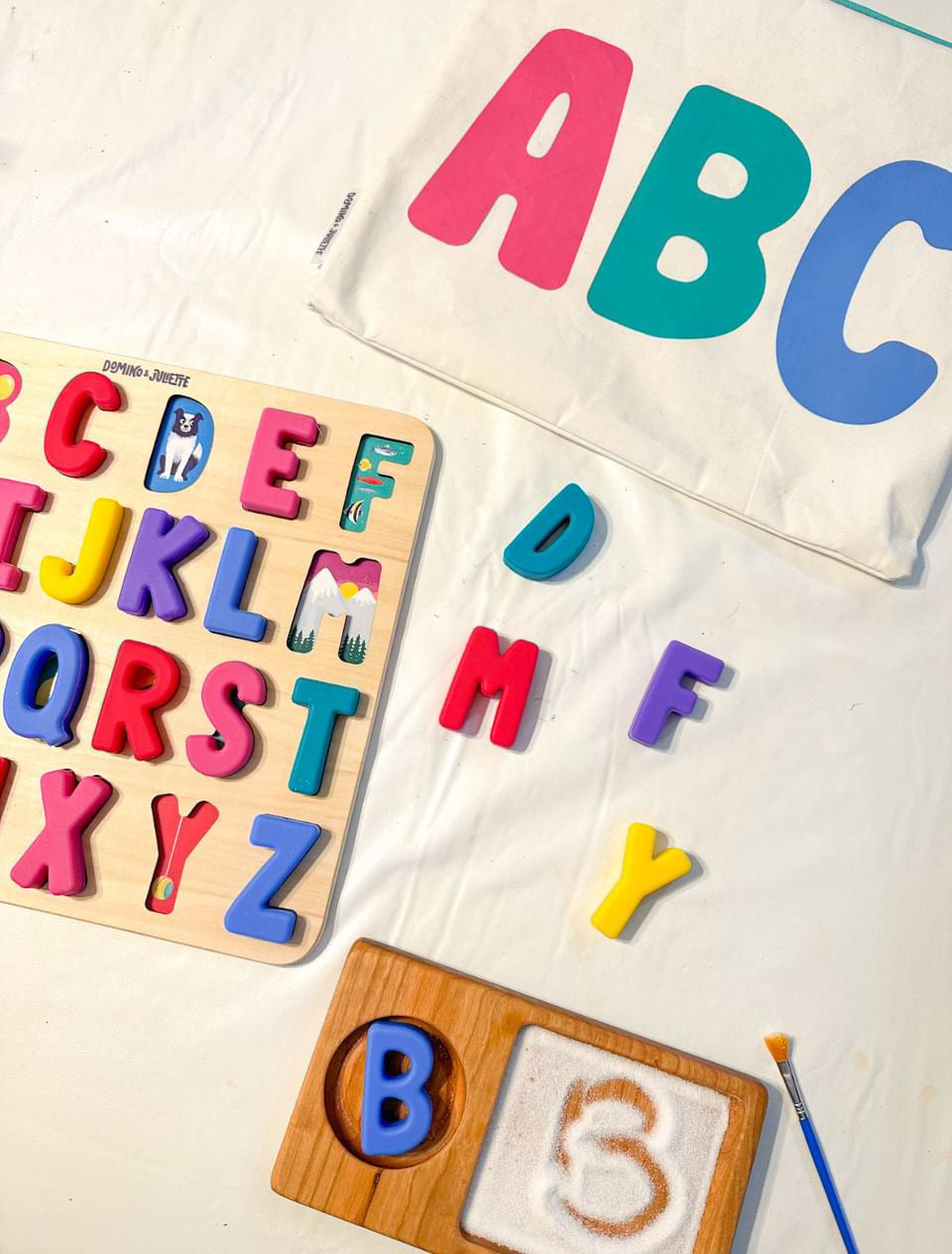 Wooden alphabet puzzle with colorful letters next to a fabric bag and a tray with letter B and textured sand for tracing