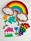 Wooden rainbow tray with white textured filler clouds, accompanied by colorful felt balls, plastic flowers, teal rain boots, butterfly shapes, and a pink house with a white fence on a white surface, for handcrafted homeschool educational resources.