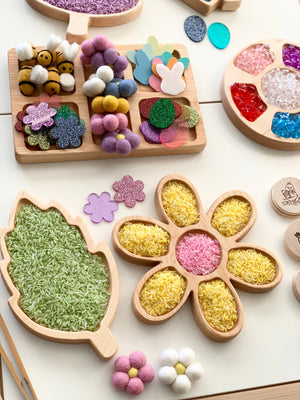 Wooden trays shaped like a leaf and flower filled with colored rice alongside felt flowers and glittery craft pieces