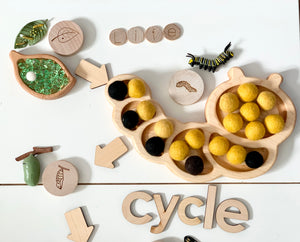 Wooden caterpillar sensory tray with yellow and black felt balls, green leaf dish, wooden letters, and insect toys on white surface