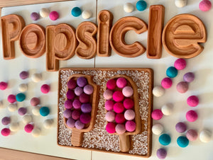 Wooden popsicle-shaped sensory tray with colorful felt balls and wooden letters spelling popsicle on white surface