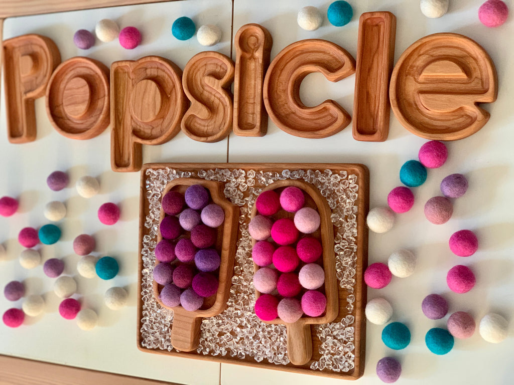Wooden popsicle-shaped sensory tray with colorful felt balls and wooden letters spelling popsicle on white surface