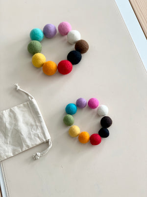 Two circles of colorful felt balls in large and small sizes with a white drawstring bag on light surface