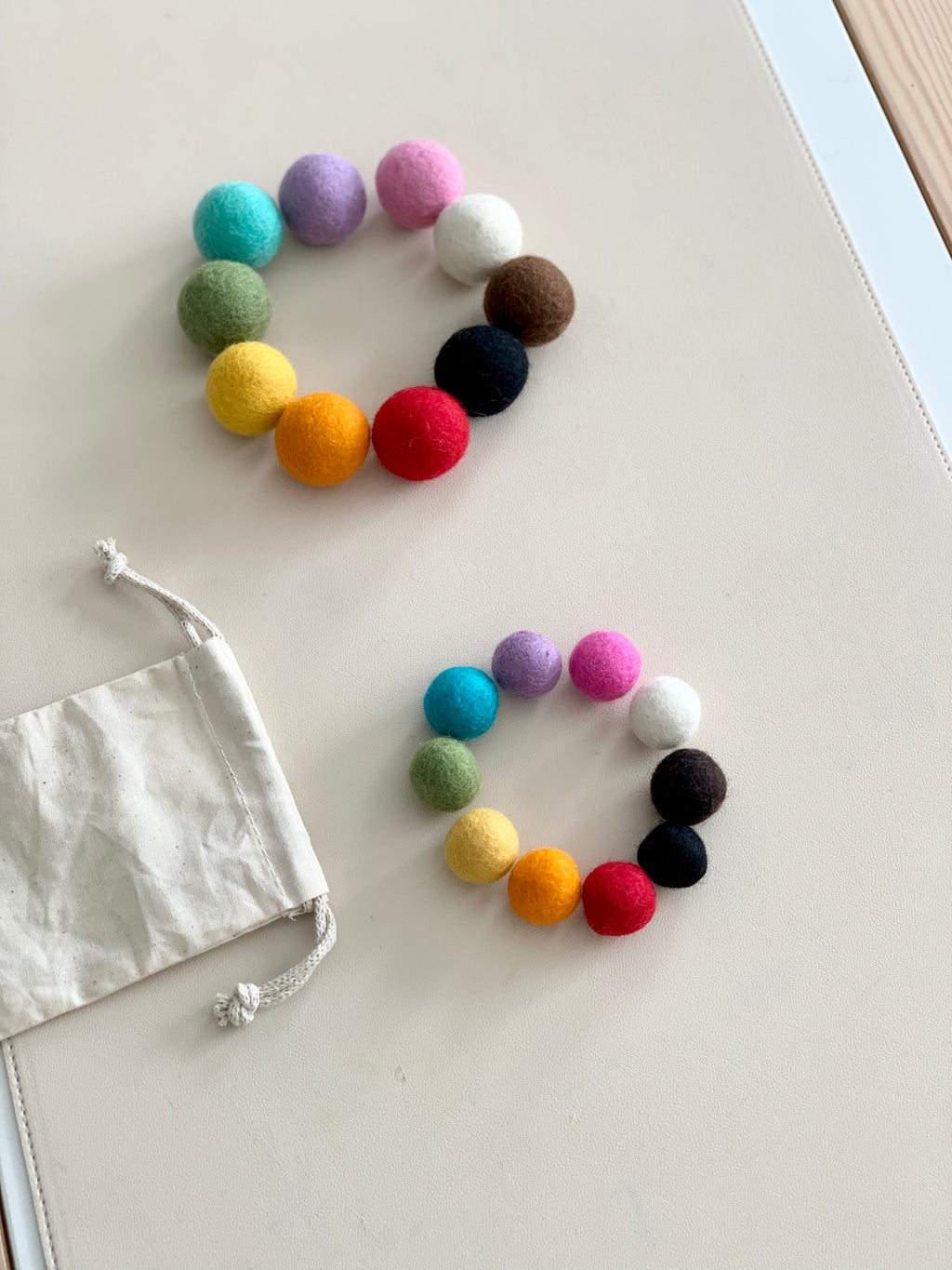 Two circles of colorful felt balls in large and small sizes with a white drawstring bag on light surface