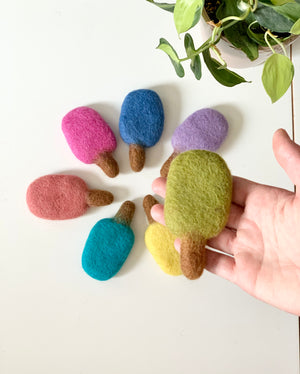 Hand holding green felt popsicle surrounded by colorful felt popsicles on white surface with green plant nearby