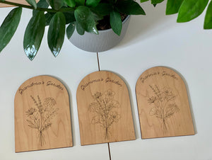 Set of three personalized wooden garden plaques engraved with wildflower bouquets labeled Grandma’s Garden on white table