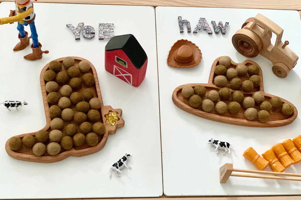 Wooden cowboy boot sensory trays filled with felt balls, small wooden farm toys, and alphabet letters on white surface