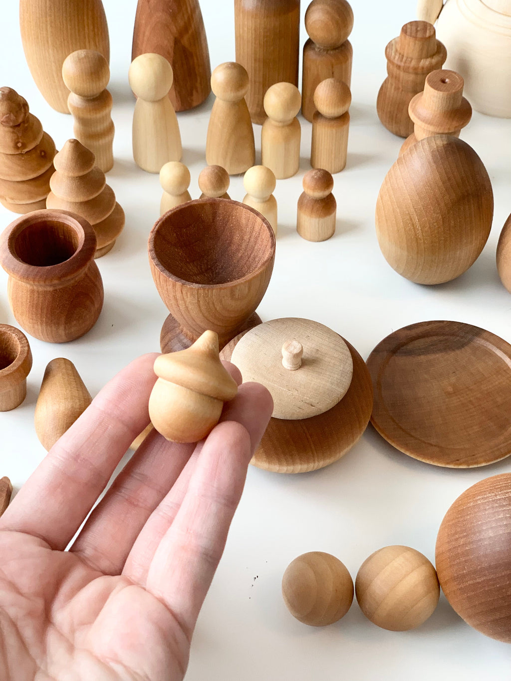 Hand holding small wooden acorn shape with various smooth unfinished wooden figures and vessels on white surface