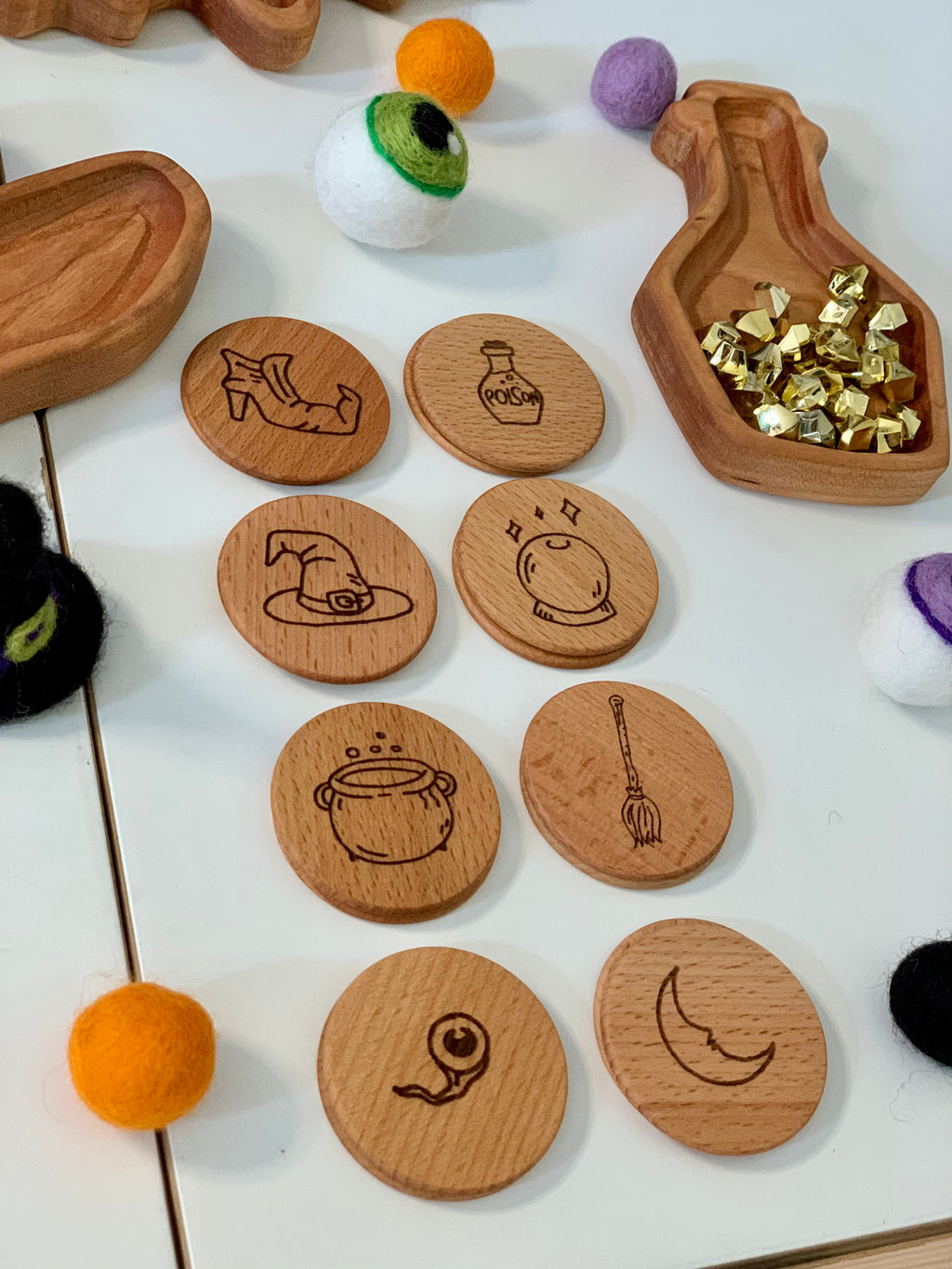 Round wooden game pieces engraved with witchy magic symbols arranged with felt balls and wooden dishes on white surface