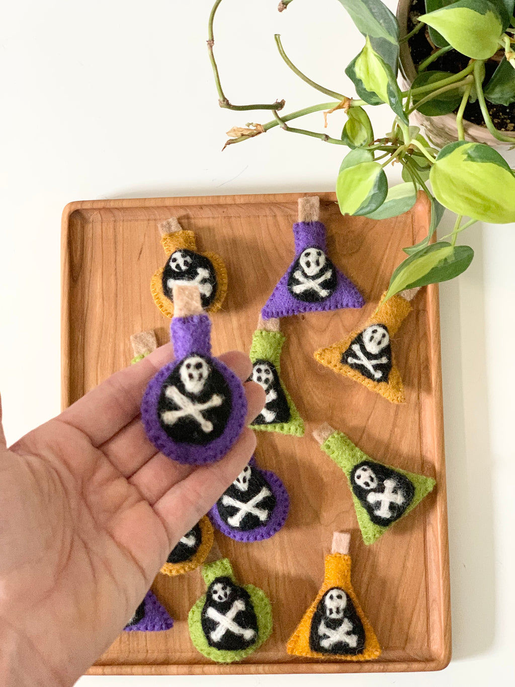 Hand holding one felt potion bottle with skull and crossbones on wooden tray with more colorful felt bottles nearby