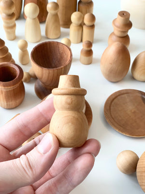 Hand holding a smooth natural wood snowman figure with assorted unfinished wooden toys and vessels on white surface