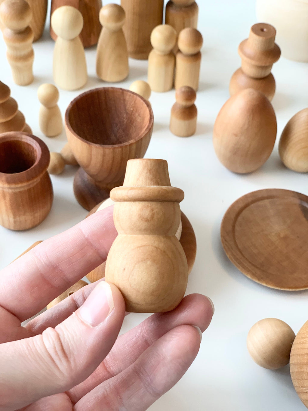 Hand holding a smooth natural wood snowman figure with assorted unfinished wooden toys and vessels on white surface