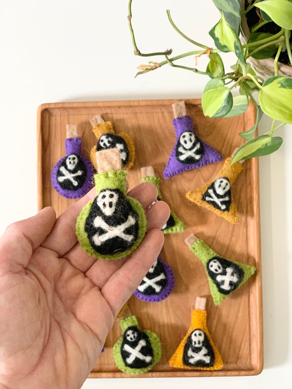 Hand holding green felt potion bottle with skull and crossbones design on wooden tray with other colorful felt bottles