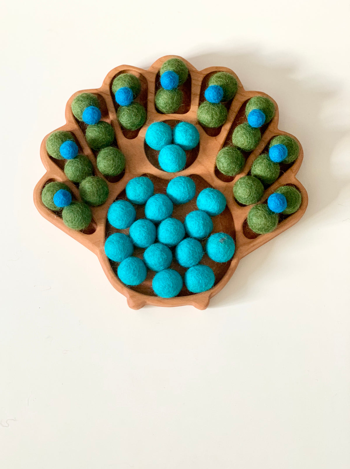 Wooden peacock-shaped sensory tray with green and blue felt balls arranged in tail feather compartments on white background