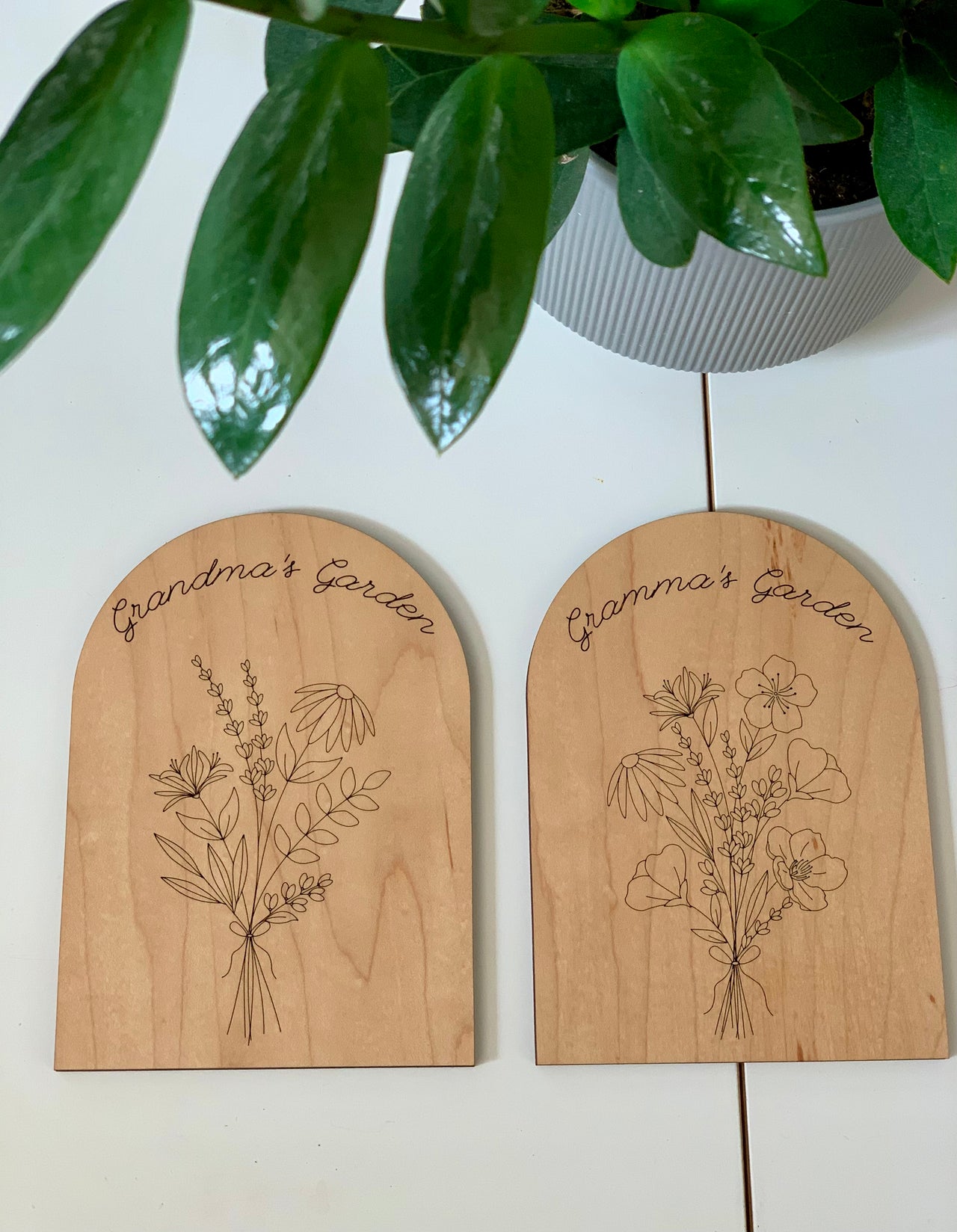 Two personalized wooden plaques engraved with floral bouquets and "Grandma’s Garden" text on white surface beneath green plant
