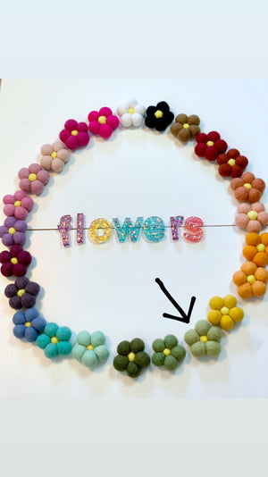 Circle of colorful handmade felt flowers with yellow centers surrounding glittery acrylic letters spelling flowers on white background