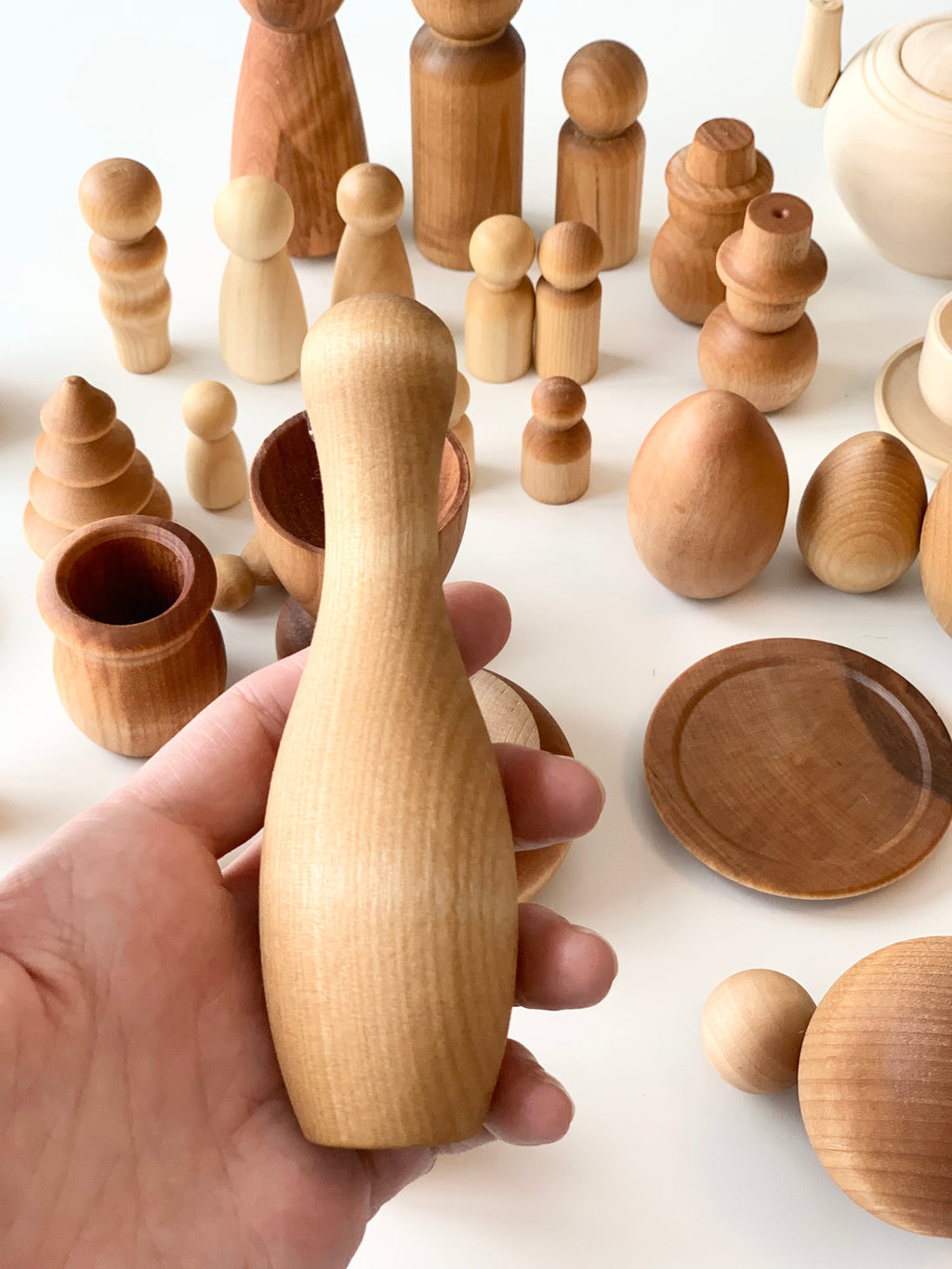 Hand holding smooth light natural wooden figure with various unfinished wooden shapes and vessels on white surface