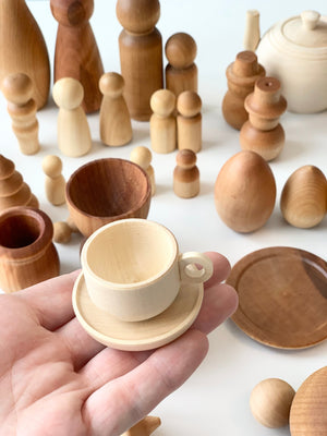 Hand holding small natural light wood teacup and saucer with an assortment of unfinished wooden toys and shapes on white surface