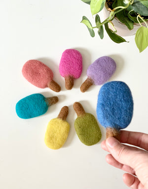 Handmade single felt popsicle in blue with additional colorful felt popsicles arranged in a circle on white surface
